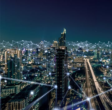 A night-time cityscape with illuminated skyscrapers and motorways. Digital white lines and glowing nodes illustrate smart city connectivity and climate action, as the bright lights stand out against the dark sky, hinting at a net zero urban future.