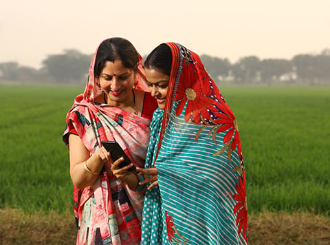 Two women in colourful sarees stand in a green field, smiling at a mobile together. The lush, blurred background highlights a rural setting where planet and people thrive in harmony with nature.