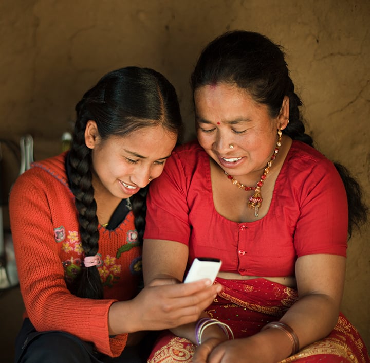Two women with dark braided hair, dressed in colorful traditional clothing, sit closely together in front of a brown wall. Smiling warmly as they look at a mobile phone, they share a joyful moment bridging the digital divide.