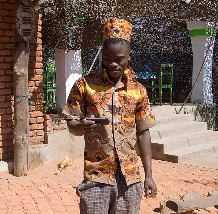 A smiling man in bright, patterned clothing and hat stands on a sunlit brick path, looking at a smartphone—bridging the digital divide in an outdoor, relaxed setting with concrete steps, shaded chairs, and foliage behind him.