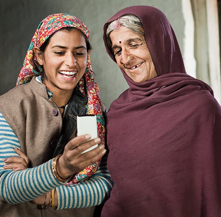 Two women wearing headscarves and traditional clothing smile and look at a smartphone together, bridging the digital divide. One is younger, in a colorful scarf and striped sweater; the other older, in a purple shawl. They share a joyful moment indoors.