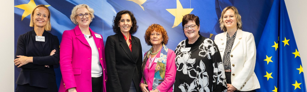 Six professionally dressed women stand smiling in front of a blue wall with large yellow EU stars and a European Union flag, attending an official event focused on International Relations within the EU, highlighting themes such as digital inclusion.