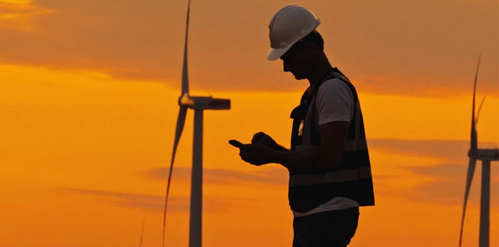 A silhouette of a worker wearing a hard hat and high-visibility vest checks a device, highlighting connectivity against an orange sunset sky. Wind turbines in the background emphasise the renewable energy setting.