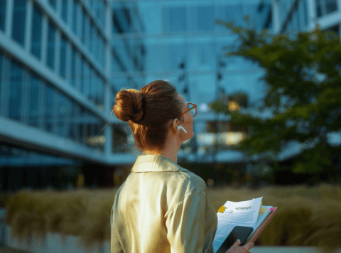 A woman with brown hair in a bun, wearing glasses and earbuds, stands outside a modern glass office building, holding documents and a phone, looking upward. Her light blazer echoes her commitment to sustainability; greenery reflects planet and people values nearby.