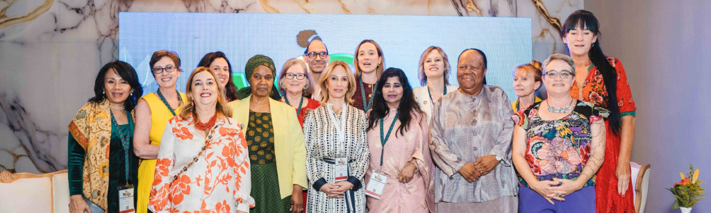 A diverse group of 16 women, some involved in Digital Inclusion and international relations, stand together smiling for a group photo against a marble-patterned backdrop at what appears to be a formal event or conference. Some wear lanyards and name badges.