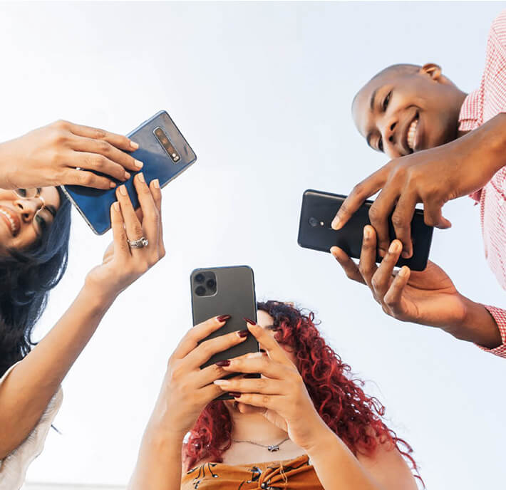 Three people stand in a circle, smiling, each holding a mobile phone and pointing them inward—perhaps exploring a case study library together. The photo is taken from below against a light sky, capturing their hands, phones, and happy faces.
