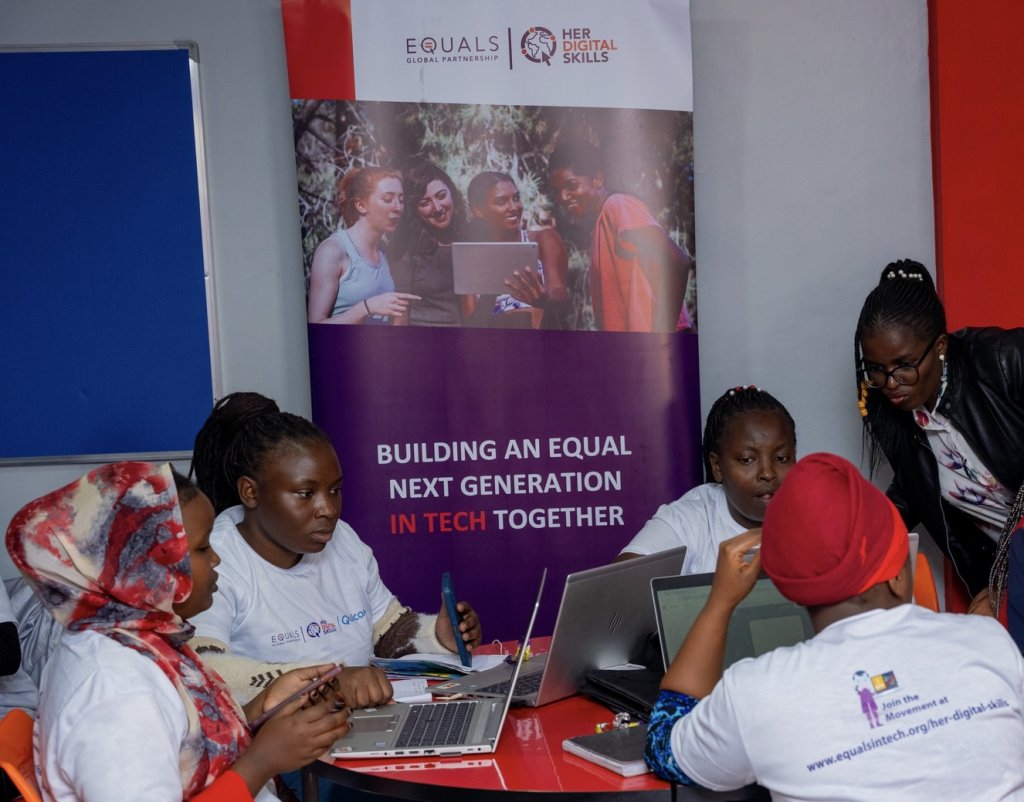 Five women collaborate around a table with laptops, papers, and pens. Behind them, a banner highlights gender diversity in tech, reading: “Building an equal next generation in tech together.” The atmosphere is focused and interactive.