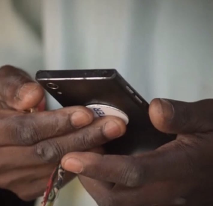Close-up of a person’s dark-skinned hands holding a black mobile phone with a round white grip on the back. The person’s thumb interacts with the phone’s screen. A multicoloured cord or strap is partially visible hanging from the phone. Background is blurred.