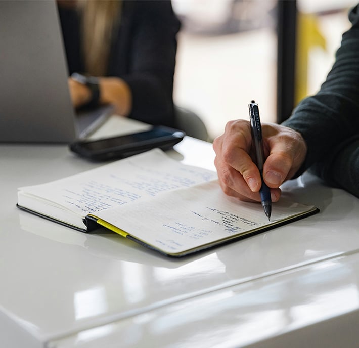 Close-up of a person’s hand writing notes in a notebook with a pen at a white desk during MWC26 Barcelona. Another person with a laptop is seated nearby, partially visible, whilst a mobile rests on the desk between them. The focus is on the workspace.