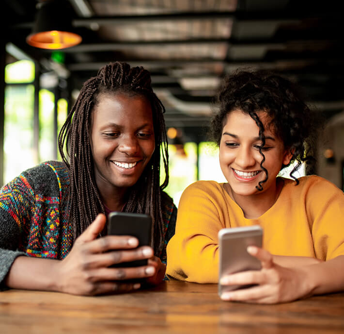 Two young women sit at a wooden table in a cosy café, smiling as they browse MWC26 Barcelona updates on their smartphones. One has plaited hair and a colourful jumper; the other, curly hair and a yellow top. Warm lighting adds to the relaxed atmosphere.