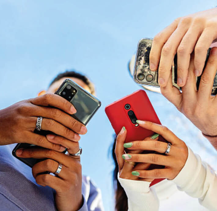 Three people stand close together at MWC26 Barcelona, holding mobile phones towards the camera. Their hands, adorned with rings, dominate the foreground whilst the blue sky fills the background, shot from a low angle focusing on phones and fingers.