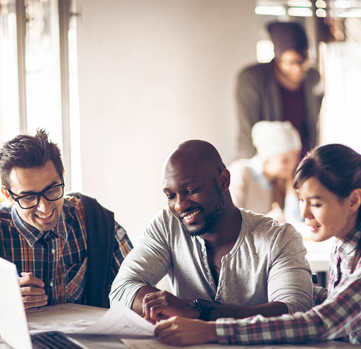Three people sit at a table, smiling and collaborating over papers and a laptop. The bright, friendly office scene captures the spirit of teamwork seen at MWC26 Barcelona, with others blurred in the background suggesting a productive environment.