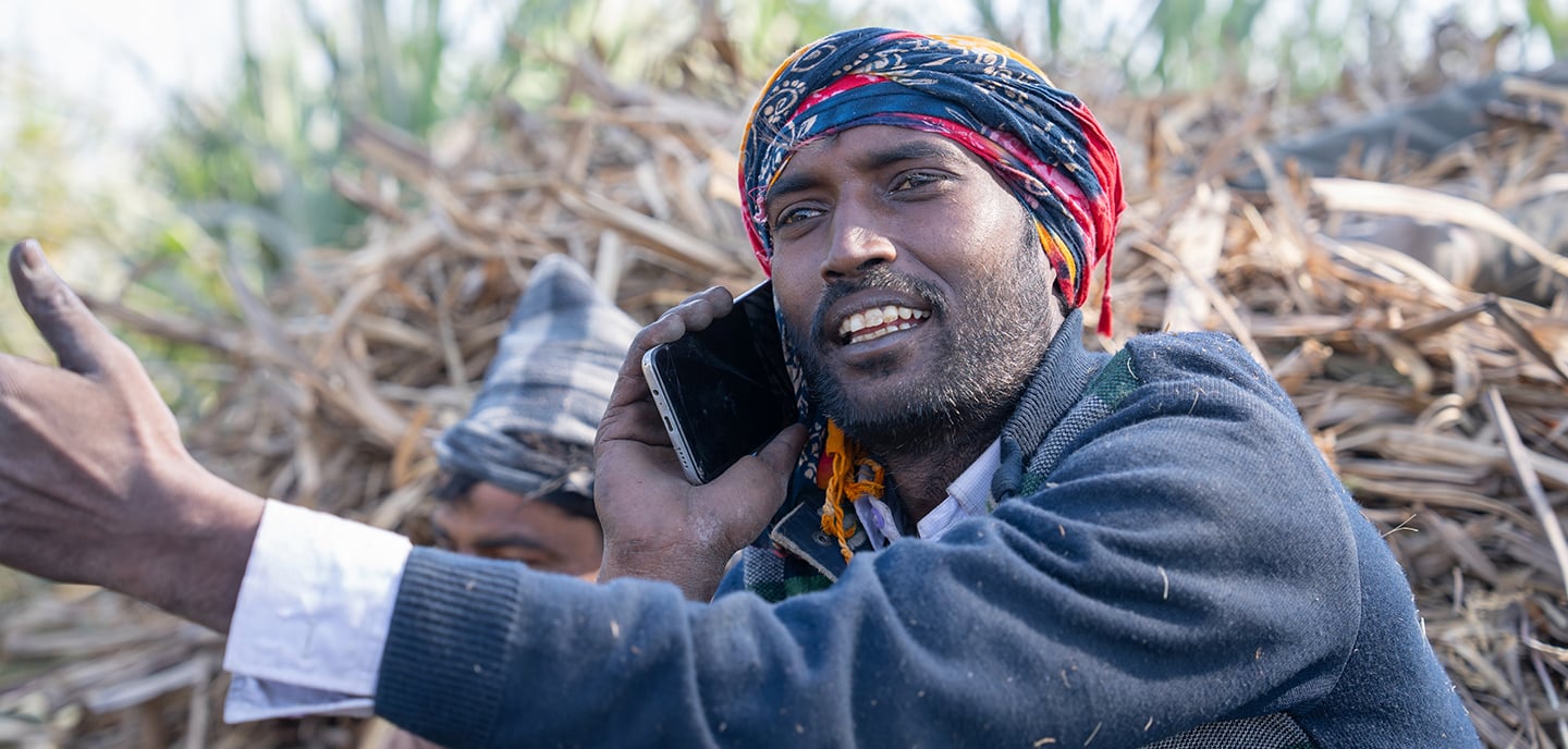 Man wearing a colourful headscarf talks on a mobile phone outdoors, sitting in front of stacked sugarcane, highlighting rural life amid climate change.