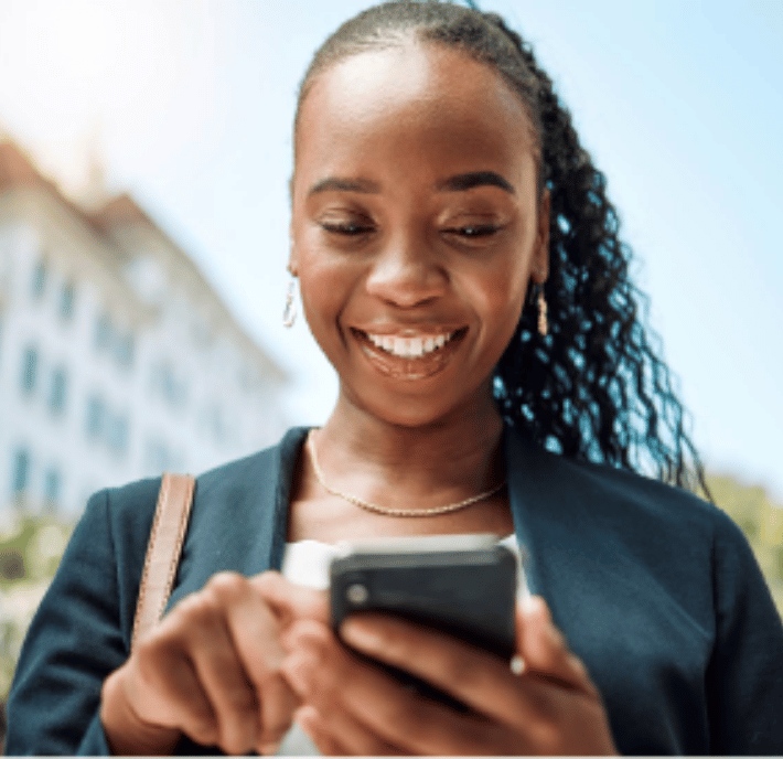 A woman with long plaited hair smiles whilst using a mobile phone outdoors. She wears a dark blazer, white top, gold necklace, and earrings. Sunlight and a blurred building appear in the background, suggesting a pleasant day.