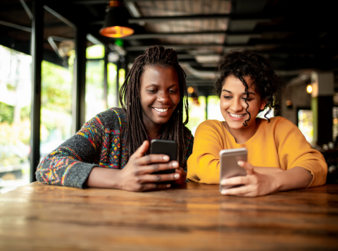 Two people sit at a wooden table in a cosy café, smiling and looking at their mobile phones. Sunlight streams through large windows behind them, and both individuals appear happy and relaxed in casual jumpers—reflecting a lifestyle that values sustainability and connection between planet and people.