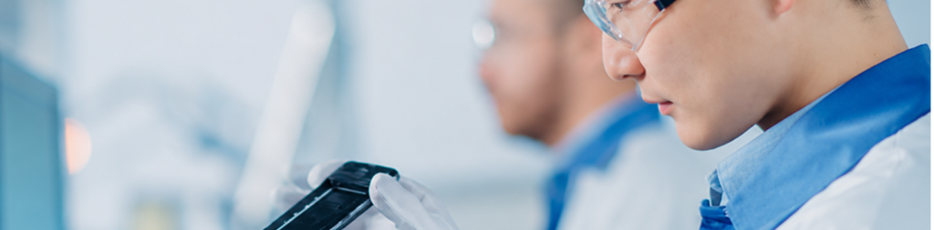 A person in safety glasses and a lab coat examines a black device whilst wearing white gloves, embodying a careful approach that considers both planet and people. Another person in similar attire is blurred in the background, suggesting a laboratory or technical work environment focused on sustainability.