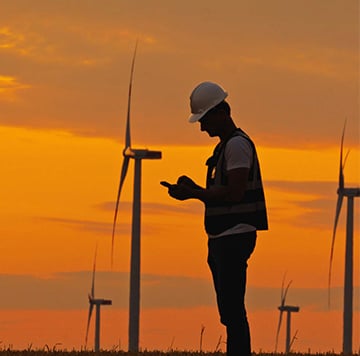 Silhouette of a worker wearing a hard hat and safety vest, standing in a field with wind turbines at sunset—a powerful image of climate action. The person appears to be using a handheld device, perhaps supporting mobile net zero initiatives. The sky is orange and yellow, with scattered clouds above the turbines.