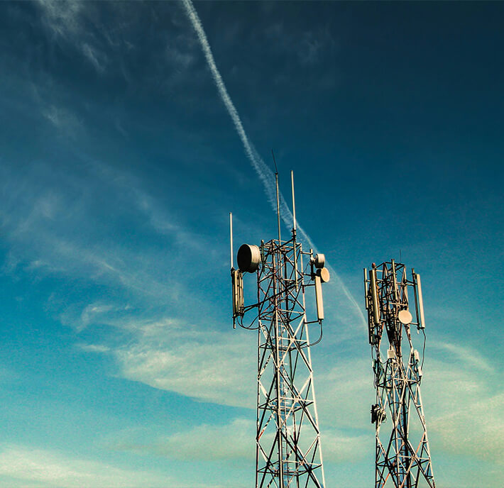 Two tall metal mobile phone masts with multiple aerials stand against a blue sky with scattered clouds, symbolising the growing connectivity across the A360 ASEAN region. A white aeroplane vapour trail diagonally crosses the sky above the masts, creating a dynamic visual element. The scene is well-lit, suggesting daytime.