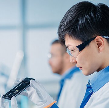A scientist in a lab coat, gloves, and safety glasses examines a black electronic component, carefully assessing its connectivity features. Another person, blurred, is visible in the background. The setting is a bright, modern laboratory.
