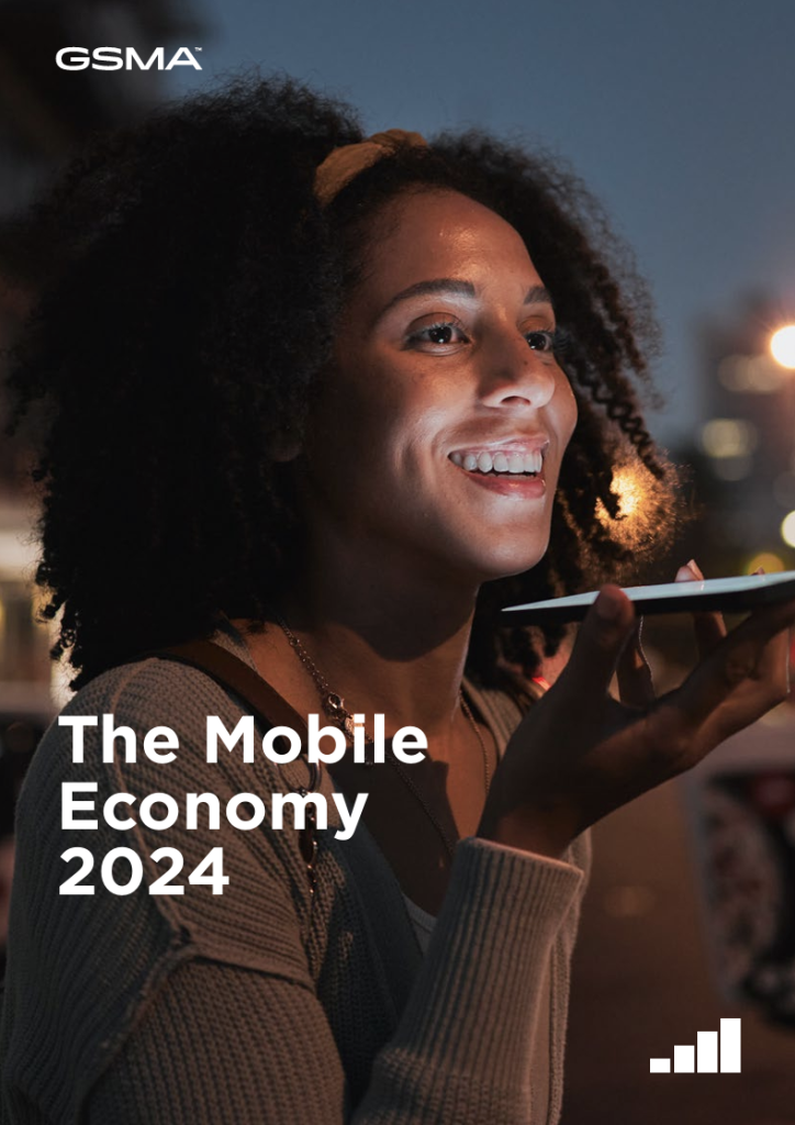 A woman with curly hair smiles whilst speaking into her mobile phone at night. City lights glow in the background. Text reads: "The Mobile Economy 2024." The GSMA logo and a signal bars icon are visible on the image.