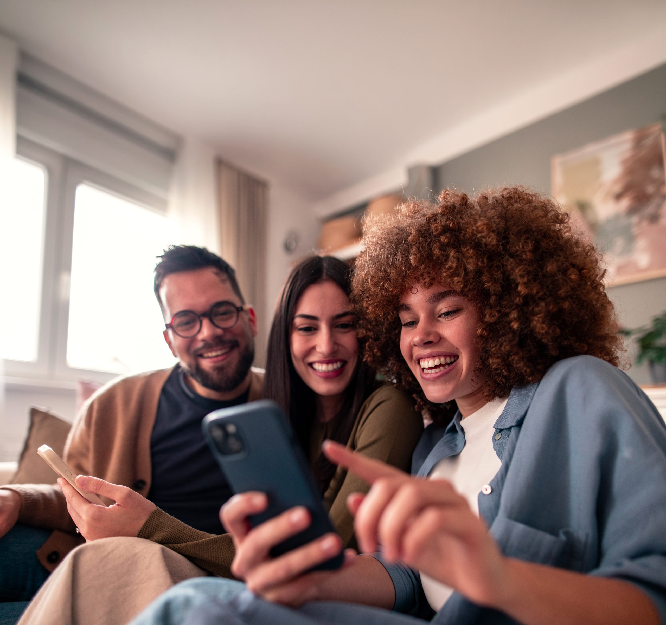 Three friends sit close together on a sofa in a bright living room, smiling and laughing whilst exploring trends in the Mobile Economy Latin America on a mobile phone. Daylight streams through the window, creating a cosy, cheerful atmosphere.