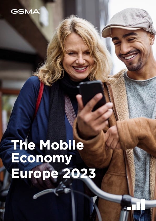 Two people, a woman with blonde hair and a man with a beard and cap, stand outdoors smiling at a mobile phone together. The woman holds the phone while the man points at the screen. Text reads: “The Mobile Economy Europe 2023.”.