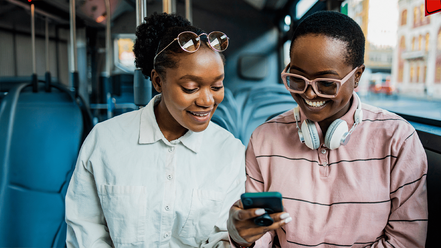 Two people sit together on a bus, smiling as they look at a mobile phone. One wears sunglasses on her head and a white shirt; the other has large glasses, headphones around her neck, and a pink striped shirt. Empty bus seats and windows are visible in the background.