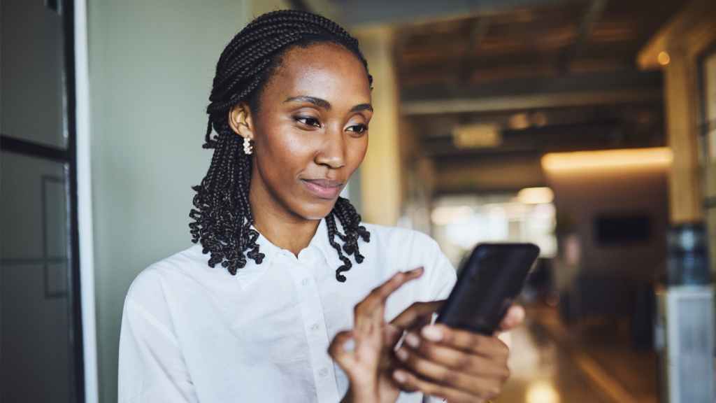 A woman with plaited hair, wearing a white blouse, looks at her mobile and gently touches the screen. She stands indoors in a softly lit modern office or workspace, with blurred background details.
