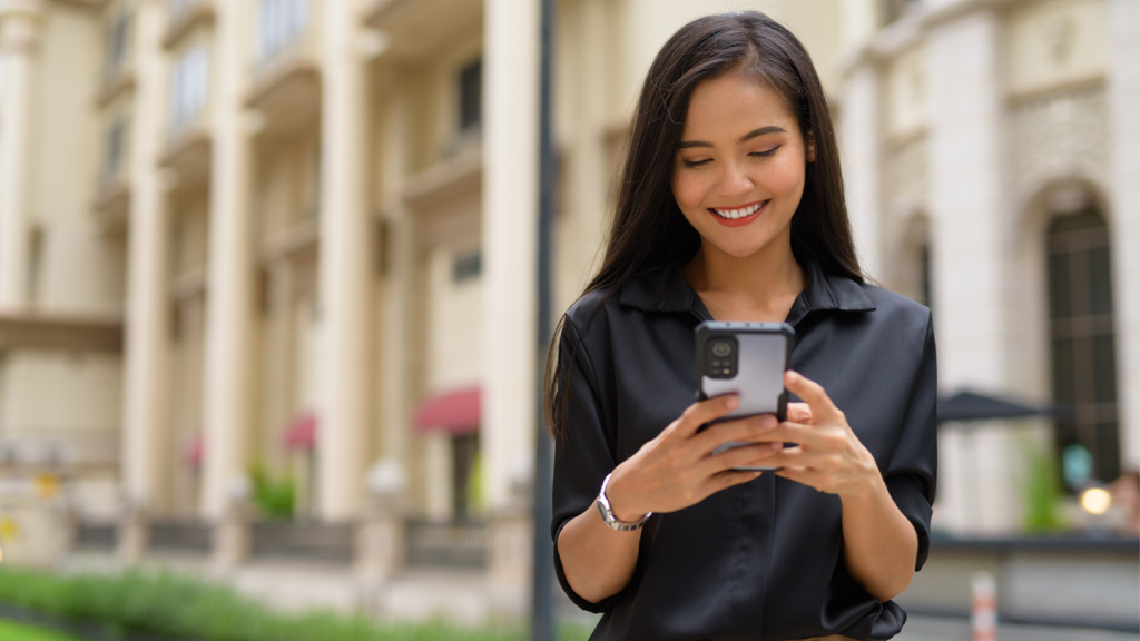 A woman with long dark hair, wearing a black blouse, smiles whilst looking at her phone outdoors. She stands in front of a beige building with columns and pink awnings. The background is softly blurred, highlighting her expression.