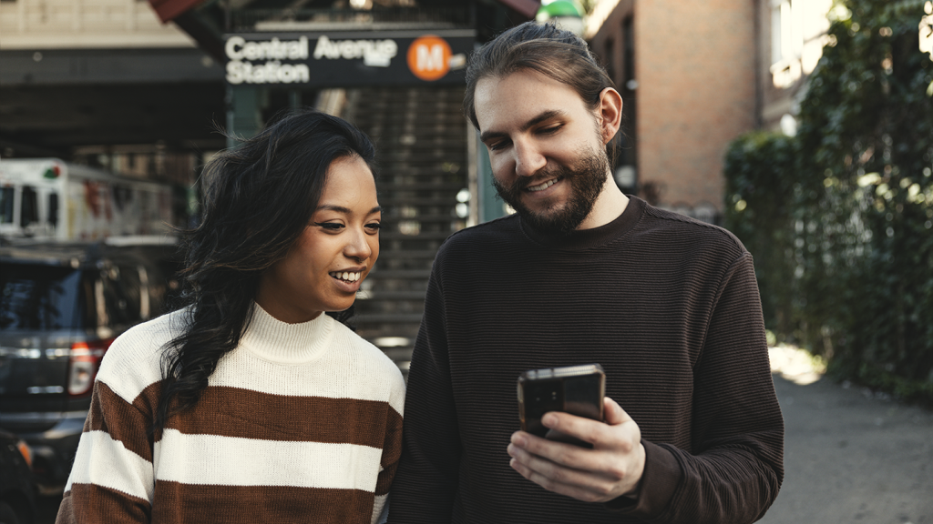 A woman and man smile whilst looking at a mobile together outdoors, standing in front of Central Avenue Station. Both wear striped jumpers. Steps and a sign are visible behind them, and cars are parked on the street.