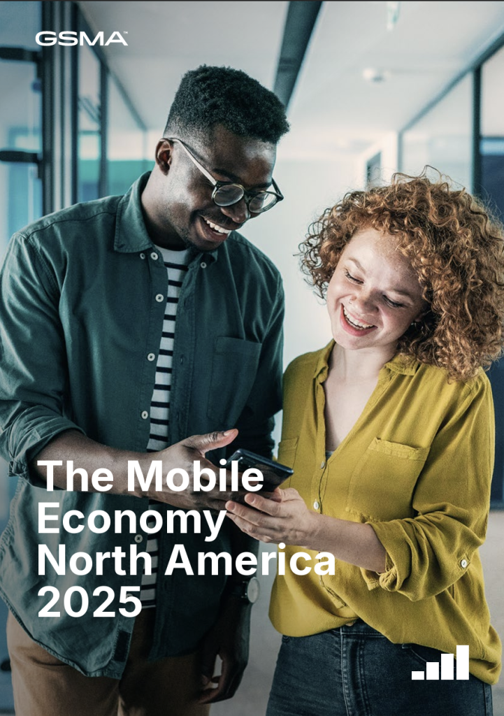Two people stand indoors, smiling whilst looking at a smartphone—capturing the spirit of the Mobile Economy North America. The person on the left wears glasses and a dark green shirt, whilst the person on the right has curly hair and a yellow shirt. Text reads: “The Mobile Economy North America 2025.”.
