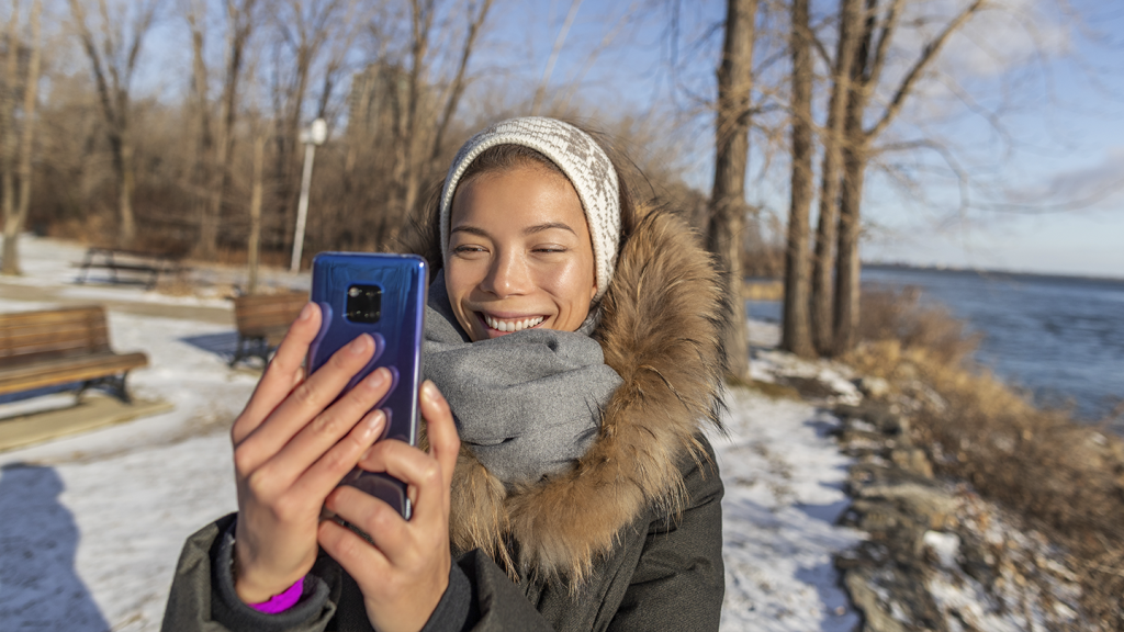 A smiling woman in a dark winter coat with a fur-trimmed hood and grey scarf takes a mobile selfie with a blue mobile phone.