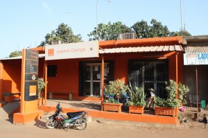 A small, bright orange building houses an "Espace Orange" shop. The shopfront has a large sign and several potted plants. A parked scooter and motorbike are in front of the entrance, and a black signpost displays the shop name and Orange logo. Trees are in the background.