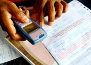 A person holding an old mobile phone in one hand while using the other hand to point at or handle paperwork and official documents spread out on a surface. The focus is on their hands, the mobile, and the documents.