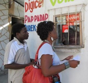 A man and woman stand outside a small building with a barred window, interacting with someone inside. The wall is painted with colourful words, including "Digicel" and "Pap Padap." The woman holds papers, and both people appear to be waiting or enquiring.