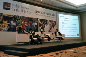 Four people sit on stage in white chairs, having a panel discussion at the "Leadership Forum 2010: Mobile Money for the Unbanked." Behind them is a large banner with photos of markets and people, plus a screen displaying forum details.