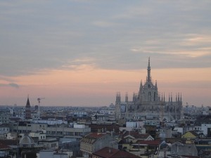 A wide cityscape view of Milan at dusk, with the iconic Milan Cathedral (Duomo di Milano) centrally visible. The cathedral’s spires rise above surrounding low buildings under a pink and blue clouded sky, suggesting the sun is setting.