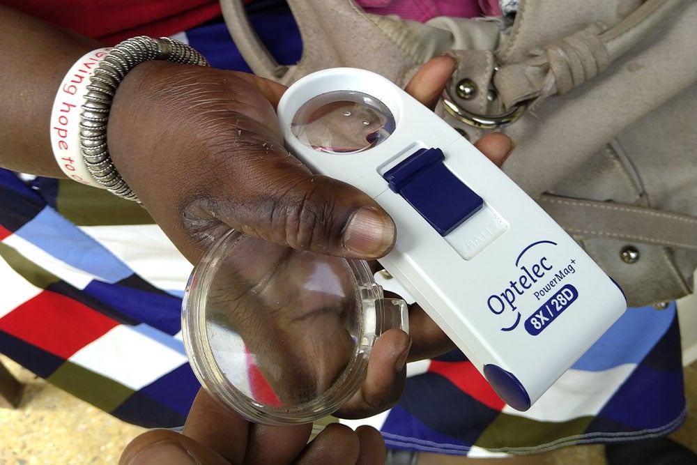 A person holds an Optelec PowerMag+ magnifying glass, labeled 8X 28D, in their right hand. They wear a patterned skirt and a silver bracelet. A wristband reads "Giving hope to." A beige bag is partially visible in the background.