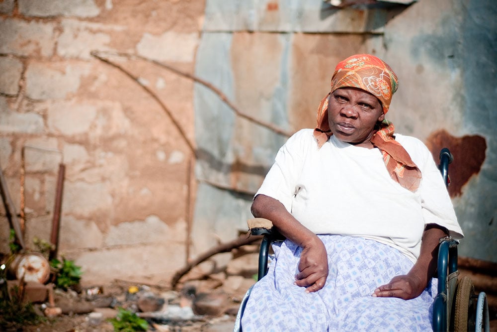 A woman in a wheelchair sits in front of a weathered brick and metal wall. She wears a white shirt, a patterned orange headscarf, and a blue patterned skirt. The background features scattered debris and natural elements, suggesting an outdoor, rural setting.