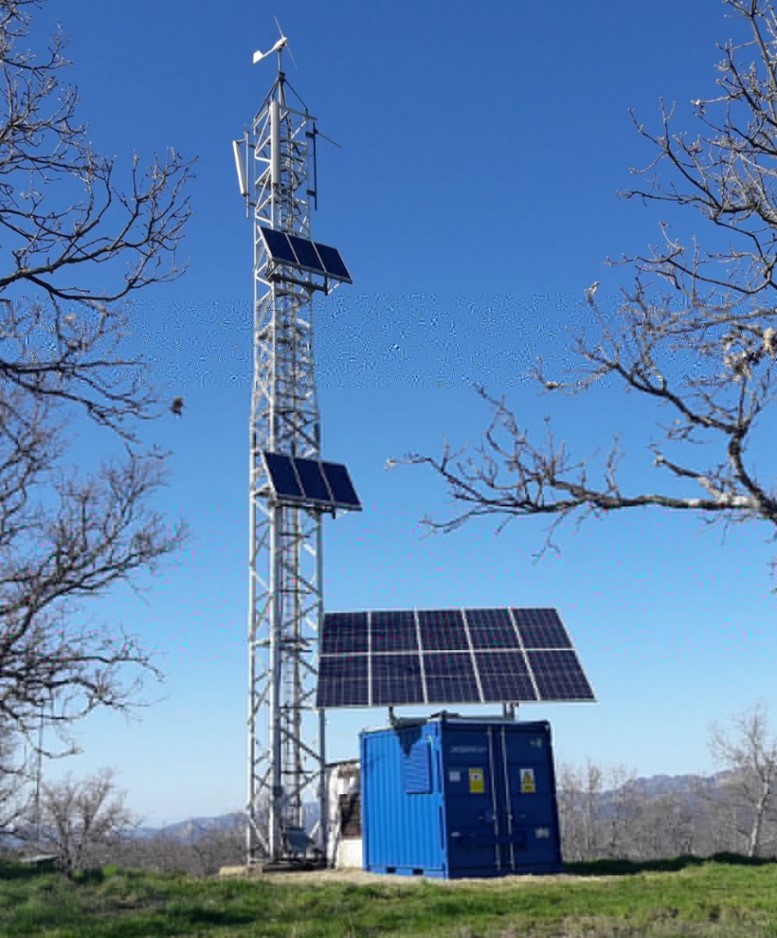 A tall metal tower with solar panels and a small wind turbine stands beside a blue container with more solar panels on its roof. Leafless trees frame the scene, under a clear blue sky, with mountains faintly visible in the background.