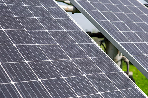Close-up of two solar panels angled towards the sun. The panels have a grid of dark photovoltaic cells with a light-colored frame. In the background, there is a blurred view of greenery, suggesting an outdoor setting.