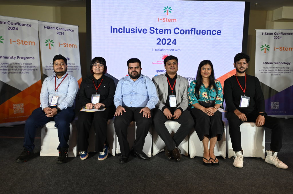 A group of six people, four men and two women, are seated side by side on a stage at the Orenda Inclusive Stem Confluence 2024 event. They are all wearing event badges. Two event banners are displayed in the background.