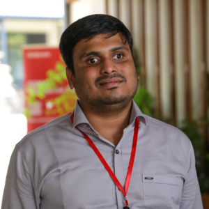 A man with short dark hair wearing a light gray button-up shirt and a red lanyard stands in a hallway with a mix of indoor and outdoor lighting. A blurred red sign and greenery are in the background.