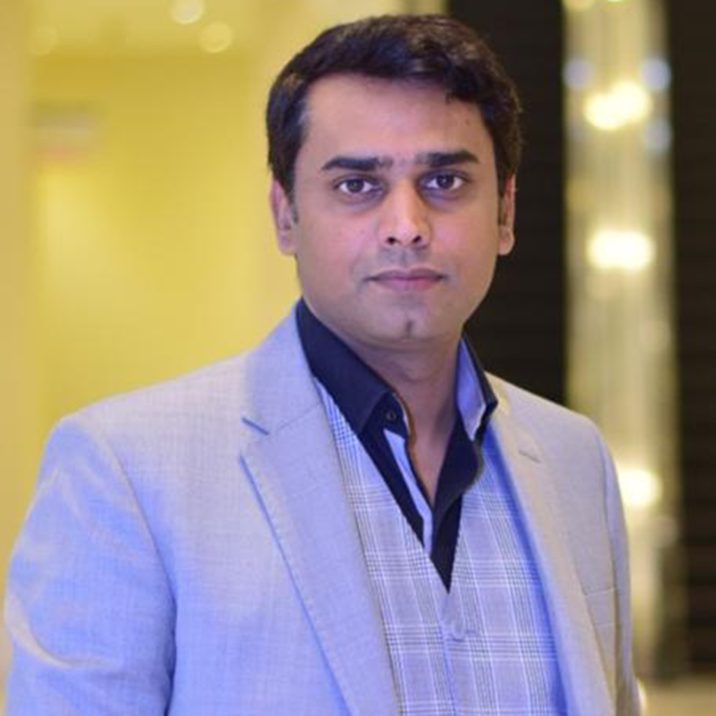 A man with short dark hair, wearing a light gray suit jacket over a gray patterned vest and dark shirt, is looking directly at the camera. The background, reminiscent of a modern knowledge platform, is blurred with warm indoor lighting.