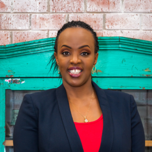 A woman with braided hair, wearing a dark blazer over a red top, stands smiling in front of a distressed turquoise door with a brick wall background. She is wearing earrings and a necklace, and her teeth are clearly visible.