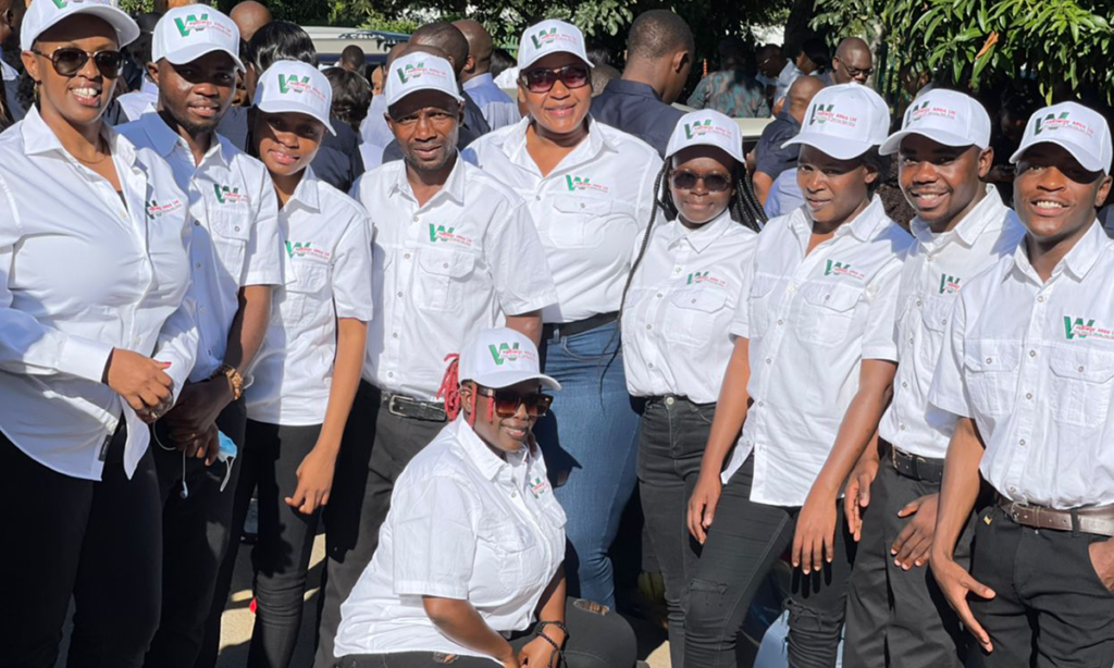 A group of eleven people poses together outdoors, all wearing white shirts and white caps with a logo. They appear to be part of a team or organization, standing in two rows, smiling at the camera. Trees and other people are visible in the background.