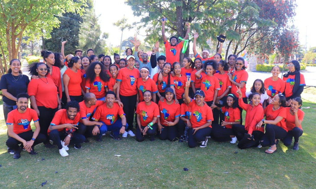 A large group of people, wearing matching red shirts with a colorful design, pose together on a grassy area under trees. Some individuals are standing while others are sitting or kneeling, with a mix of smiling and cheering expressions—all proudly representing Africa 118.