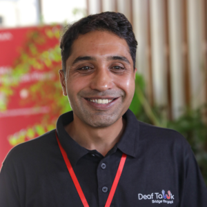 A smiling man wearing a black polo shirt with a "Deaf to UK" logo, standing indoors with a blurred background featuring a red banner.