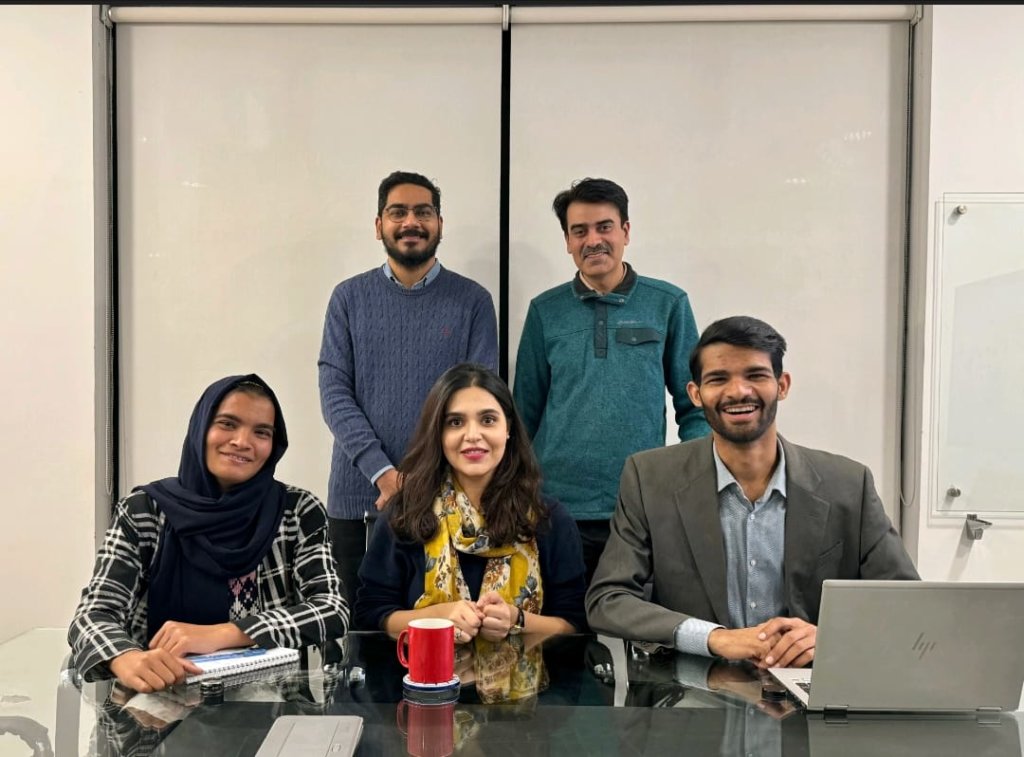 A group of five people is in a meeting room. Three are sitting at a table, with a woman in the center and a man and woman on either side. Two men are standing behind them. The table has a red mug, a laptop, a notebook, and some papers.