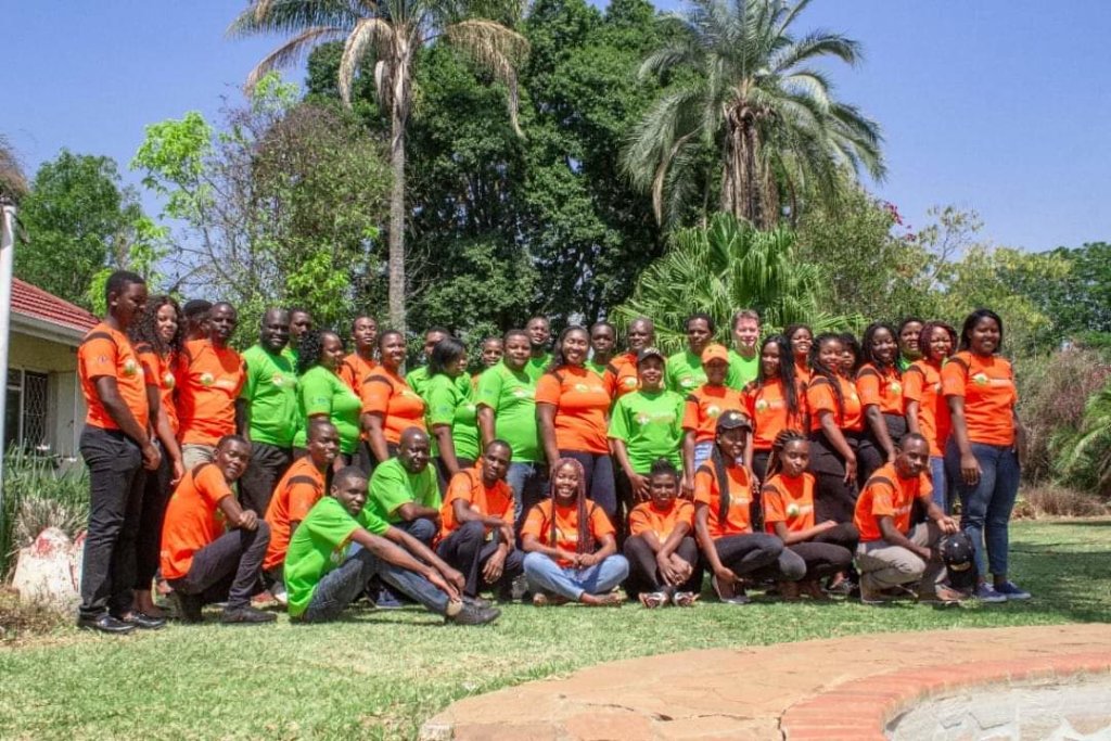A large group of people, wearing orange and green shirts, are gathered outdoors on a sunny day, radiating Zonful energy. Some are standing while others kneel or sit on the grass. A variety of trees and a clear blue sky can be seen in the background.